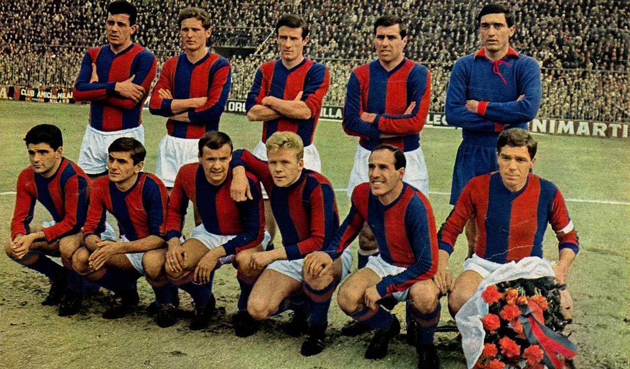 Eleven players of the 1963-64 Bologna Football Club pose for a team photo on the pitch in front of a crowded stadium. Ten players wear red and blue striped jerseys and white shorts, while the goalkeeper wears an all-blue kit. A player in the front row holds a bouquet of red flowers.