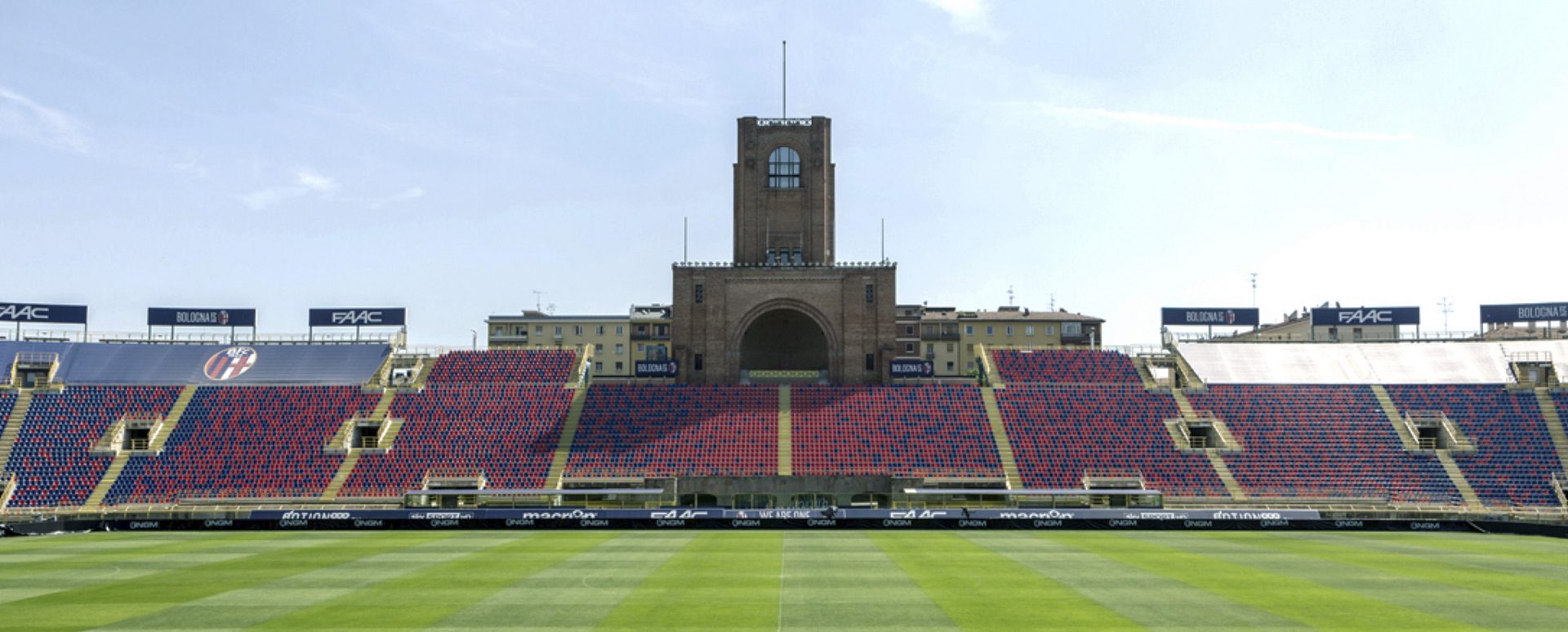 An empty football stadium featuring stands with alternating red and blue seats, a green pitch in the foreground, and a distinctive brick tower structure with an archway rising behind the seating area.
